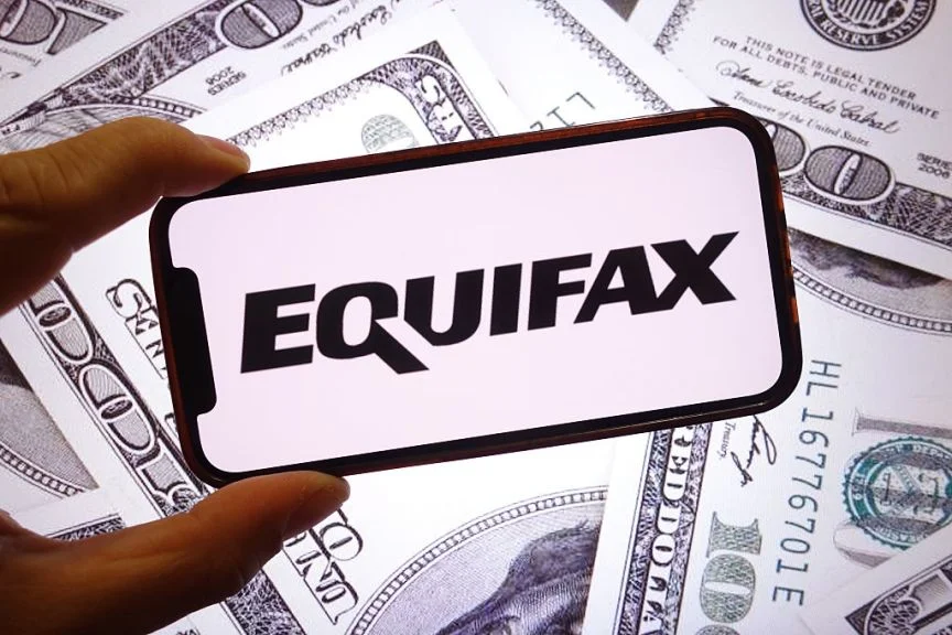 A hand holds up a phone with a black Equifax logo plastered on it. US dollars are scattered around the background.