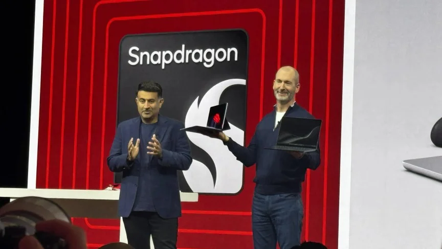 Two men stand on stage, one clapping while another holds two laptops up.