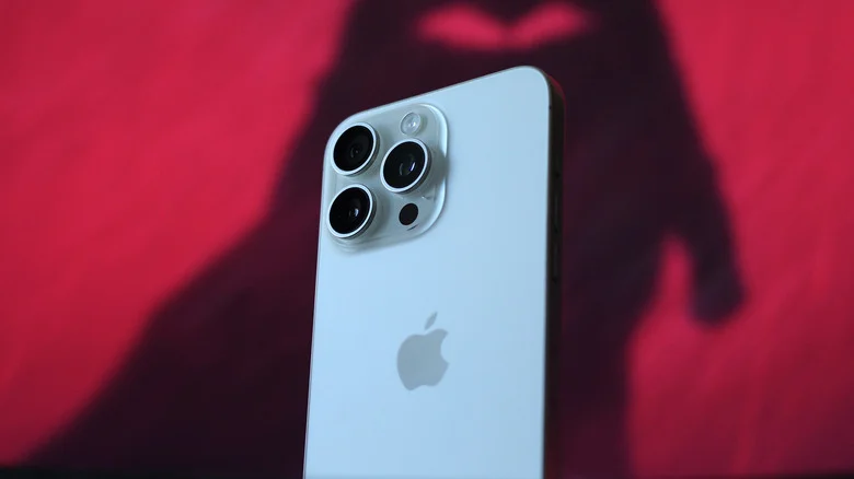 The back of a white iPhone 15 Pro Max, with a shadow on the red wall behind it.